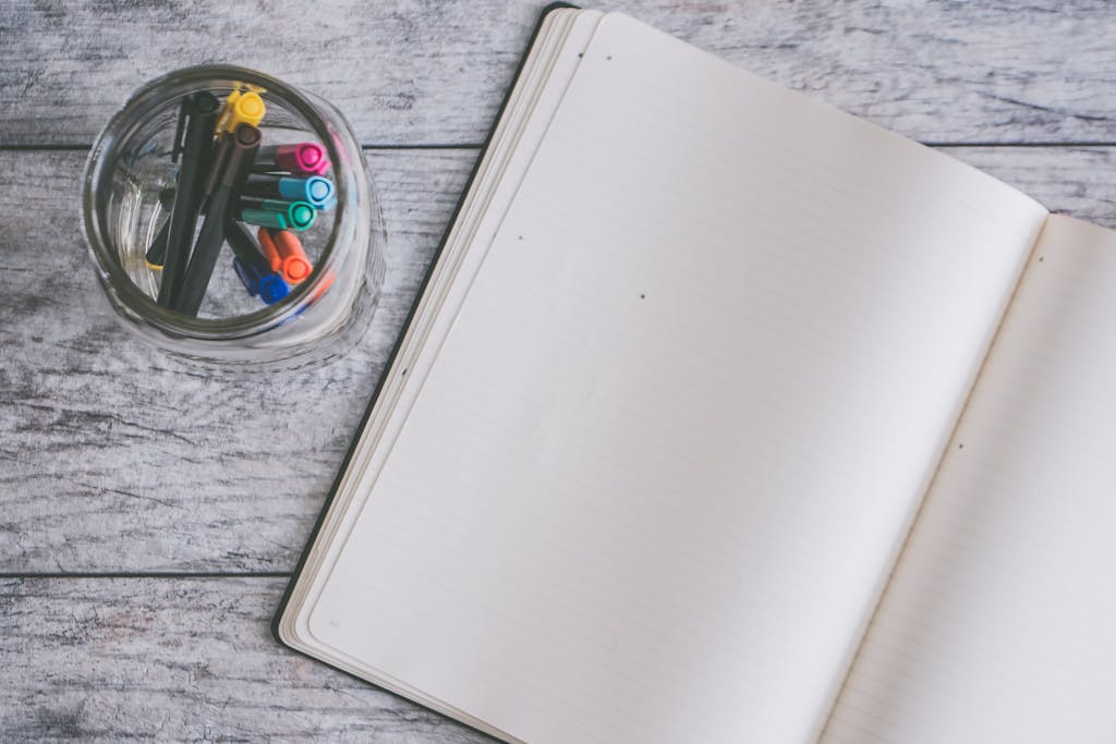 An open notebook and colorful pens in a glass jar on a wooden desk.